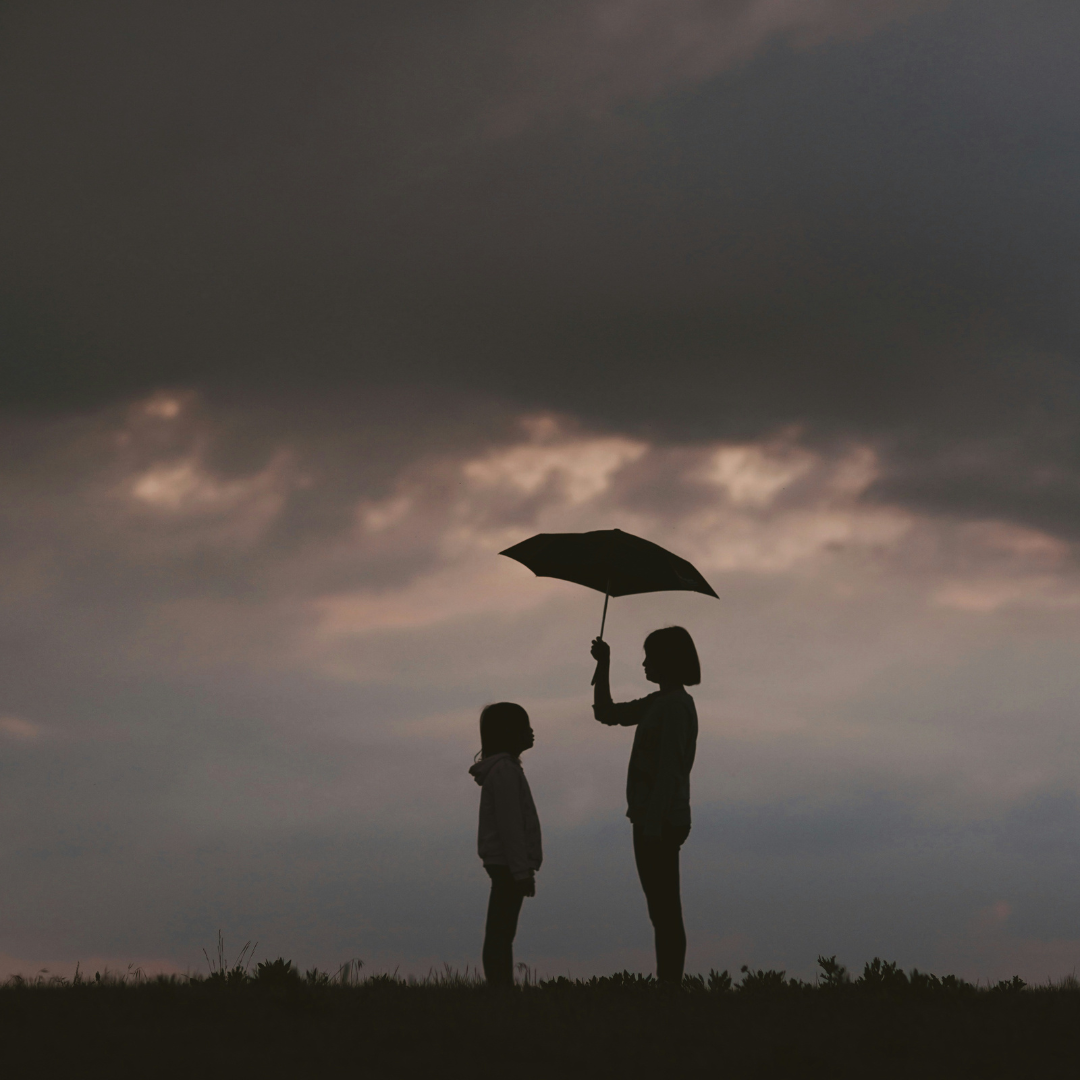 Silhouette d’une personne tenant un parapluie pour en protéger une autre sous un ciel sombre, symbole de soutien et de solidarité.